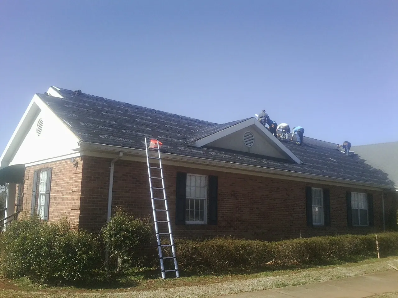 Crew tearing shingles off a brick ranch roof with a ladder set — Metal Roof Repair work by our crew in Fountain Inn