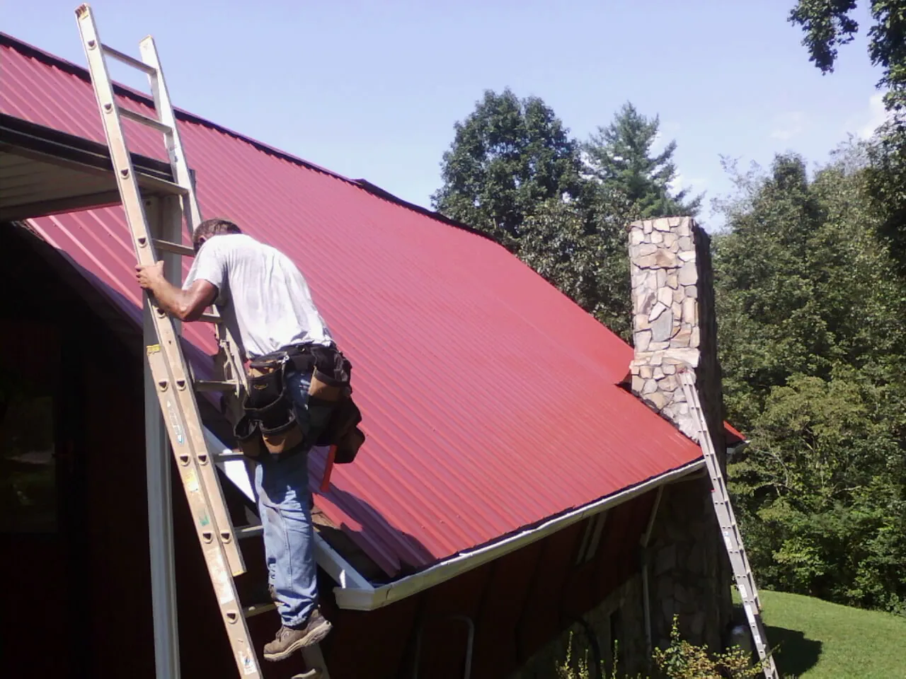 Roofer on a ladder beside a red metal roof and stone chimney — Soffit & Fascia Repair by Roofing Brothers in Fountain Inn